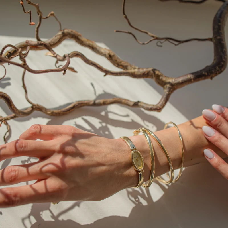 Dłoń kobiety z białym manicurem prezentująca złote bransolety i złoty zegarek na nadgarstku. Dłoń leży na jasnym tle z ciekawymi cieniami gałęzi, które dodają artystycznego nastroju zdjęciu.