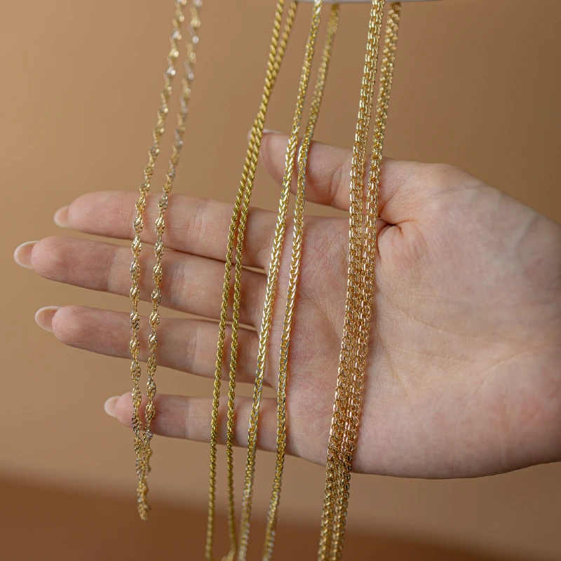 Dłoń prezentująca kilka różnych wzorów złotych łańcuszków na neutralnym, beżowym tle.