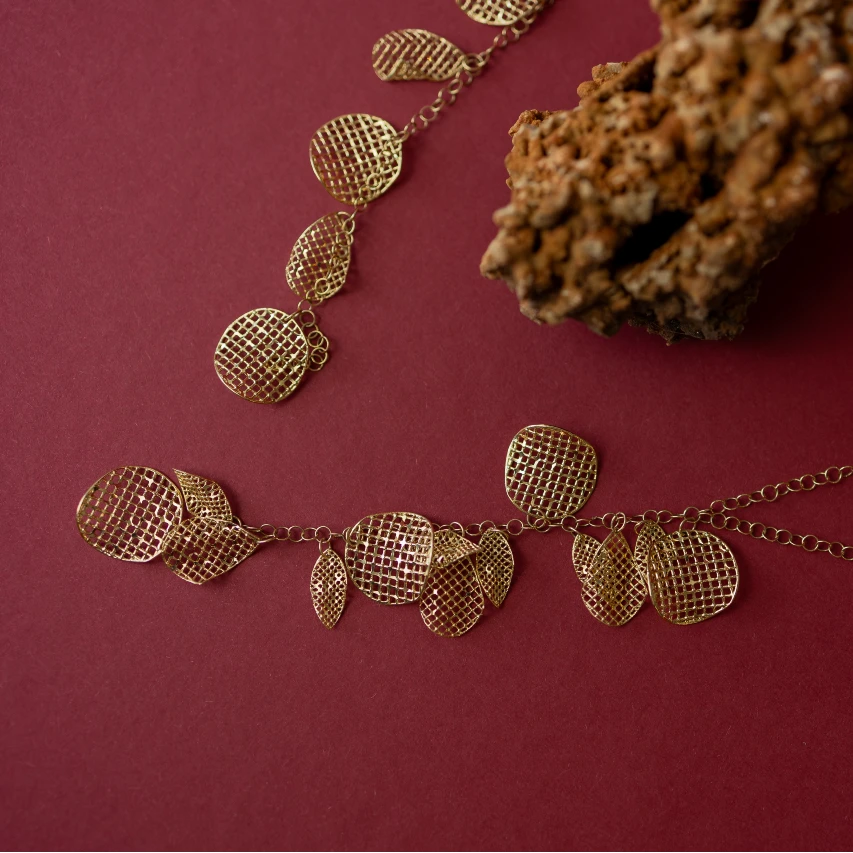 Elegancki złoty naszyjnik z wieloma ażurowymi, siatkowymi elementami o różnych kształtach, rozłożonymi symetrycznie na ciemnoczerwonym tle. Biżuteria prezentuje intrygujące połączenie okrągłych i owalnych oczek, które tworzą przyciągający wzrok wzór, dodając uroku i subtelności całościowej kompozycji.